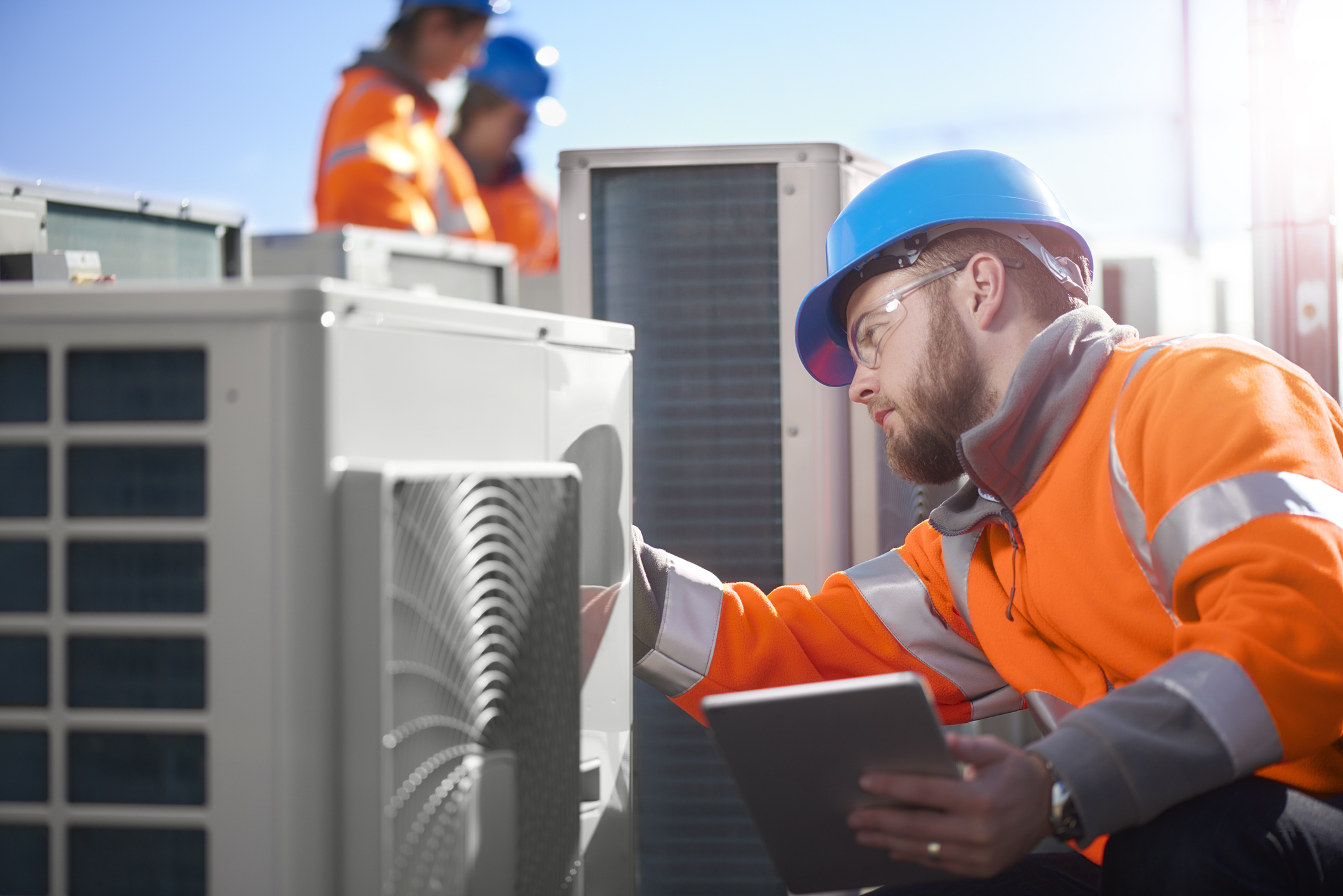 An air conditioning engineer is finishing the installation of several units on a rooftop. Two colleagues can be seen also installing units in the background. They are wearing hi vis jackets, hard hats and safety goggles.